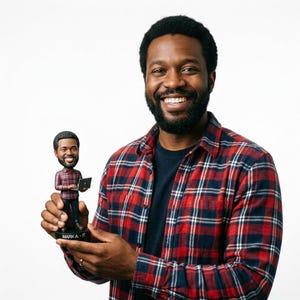 May include: A bobblehead figurine of a man holding a laptop, wearing a red and black plaid shirt, and blue pants. The figurine has the text "MARKA - IT" on the base. The man in the image is also wearing a red and black plaid shirt.