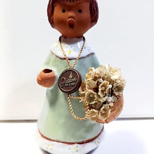 May include: A ceramic figurine of a person with brown hair wearing a light green dress with white trim and yellow dots. The figure holds a brown basket filled with beige flowers and wears a gold cord necklace with a round brown Jallar Made in Spain tag.
