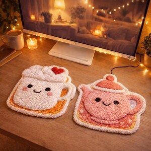 May include: Two plush, decorative coasters. One is shaped like a coffee cup with whipped cream and a red heart. The other is a pink teapot. Both have smiling faces and are on orange-yellow mats.