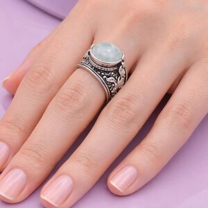 Pode incluir: Um anel de prata com uma pedra preciosa ovalada verde claro. O anel tem um design ornamentado e detalhado ao redor da banda e da cravação da pedra preciosa. O anel está em um dedo, repousando sobre uma superfície roxa.