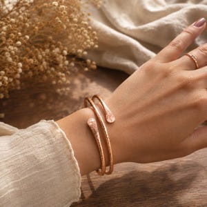 Peut inclure: Un bracelet manchette couleur or rose présentant un design à double enroulement avec une finition martelée texturée. Le bracelet possède deux extrémités arrondies et aplaties avec un éclat métallique, porté au poignet avec une bague simple.