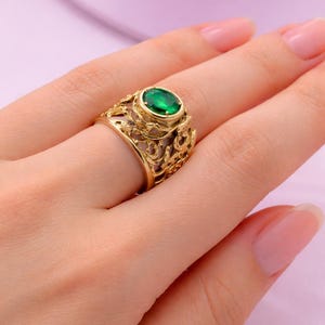 Pode incluir: Um anel dourado com uma pedra preciosa oval verde esmeralda. O anel apresenta um design ornamentado e vazado com padrões em espiral. O anel está em um dedo, mostrando os detalhes intrincados e a cor vibrante da joia.