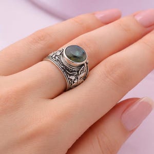 Pode incluir: Um anel de prata com uma pedra preciosa oval verde escuro. O anel tem um design floral ornamentado ao redor da banda e da configuração da pedra. O anel está em um dedo.