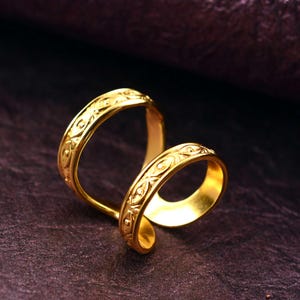 Puede incluir: Primer plano de un anillo dorado con un patrón intrincado y repetitivo. El anillo tiene un diseño único, con dos bandas que se curvan y se superponen. El anillo está hecho de un material metálico brillante, y los detalles del patrón son claramente visibles.