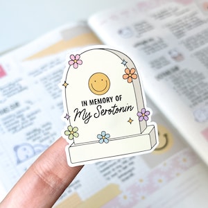 Könnte beinhalten: Ein Aufkleber in Form eines Grabsteins mit der Aufschrift "IN MEMORY OF My Serotonin". Der Grabstein hat ein gelbes Smiley-Gesicht und ist mit bunten Blumen und Sternen verziert. Der Aufkleber wird von einem Finger gehalten.