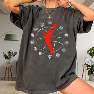 Puede incluir: Camiseta gris oscuro con una silueta roja de una jugadora de baloncesto alcanzando una pelota, rodeada por un diseño de baloncesto blanco y logotipos de equipos. La camiseta tiene un aspecto vintage y desgastado.