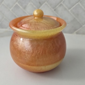 May include: A small, round, lidded jar with a warm, shimmering color palette. The jar is primarily a blend of orange and gold, with a rounded body and a lid topped with a small knob. The rim and knob have a golden hue.