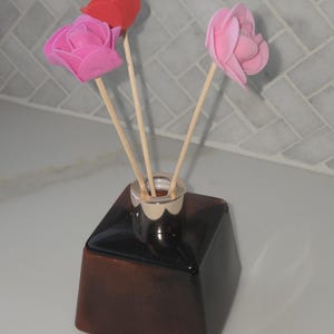 May include: A brown square diffuser bottle with a silver collar holds three faux rose flowers. The roses are pink and red, and are attached to wooden sticks. The diffuser sits on a white surface.