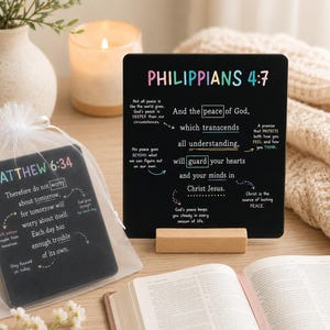 May include: Two black square plaques featuring colorful text and white scripture verses from Philippians 4:7 and Matthew 6:34. One plaque stands on a light wooden base, while the other is inside a white sheer drawstring bag on a wooden surface.