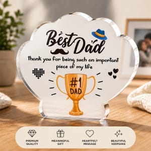 Puede incluir: Un recuerdo de acrílico transparente con las palabras "Best Dad" y "Thank you for being such an important piece of my life." Presenta un bigote, un sombrero azul y un trofeo dorado que dice "#1 Dad."