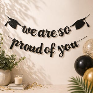 Könnte beinhalten: Ein schwarzes Glitzerbanner mit den Worten "we are so proud of you" wird angezeigt. Zwei Abschlusskappen befinden sich auf dem Banner. Ballons in Gold, Schwarz und Transparent mit goldenen Konfetti befinden sich im Hintergrund. Eine Kerze und Grünzeug befinden sich im Vordergrund.