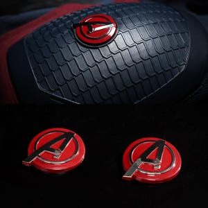 May include: A close-up of a navy blue and red costume detail with a textured, scale-like pattern. The design includes a red circular emblem with a silver "A" and a red circle with a silver arrow. Two additional red and silver "A" emblems are also visible.