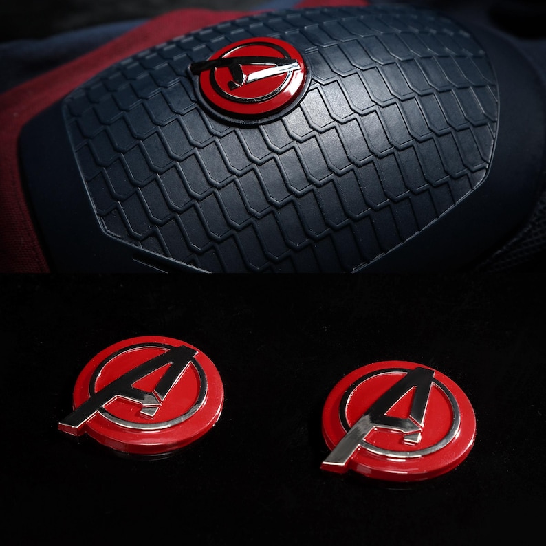 May include: A close-up of a navy blue and red costume detail with a textured, scale-like pattern. The design includes a red circular emblem with a silver "A" and a red circle with a silver arrow. Two additional red and silver "A" emblems are also visible.