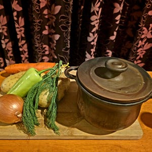 May include: A brown ceramic cooking pot with a lid and handles rests on a wooden board. Beside it are fresh vegetables: a carrot, green pepper, onion, potatoes, and dill. The scene suggests a culinary theme, perfect for a home kitchen.