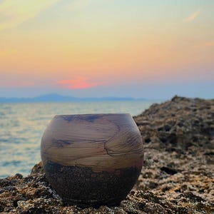 May include: A round ceramic vase with a textured, earthy brown and dark brown finish. The vase is set on a rocky surface with the sea and a colourful sunset in the background. The vase has a unique, organic shape.