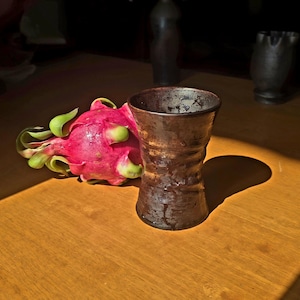 Puede incluir: Un jarrón de cerámica marrón con superficie texturizada se encuentra junto a una vibrante fruta del dragón rosa con hojas verdes. El jarrón tiene una abertura ancha y una base ligeramente ensanchada. Los objetos están sobre una superficie de madera.