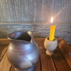 Puede incluir: Una jarra de cerámica azul oscuro hecha a mano con un pico, junto a una vela amarilla encendida en un pequeño soporte de color claro. Los objetos están sobre una superficie de madera, con un fondo gris oscuro.