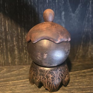 Puede incluir: Un tarro de cerámica marrón oscuro hecho a mano, con tapa y base decorativa. El tarro tiene un cuerpo redondeado y una tapa con un pequeño pomo. La base tiene un patrón texturizado único. El diseño general es rústico y terroso.