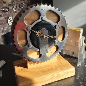 May include: A distinctive clock crafted from a motorcycle sprocket, with a silver gear displaying the number 45, mounted on a wooden backing and base. The clock hands are gold, with a red second hand. A decorative item.