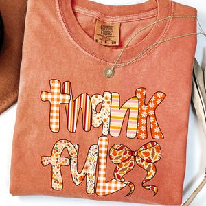 May include: A terracotta-coloured short-sleeve t-shirt featuring the word "thankful" in a multi-patterned graphic print. The letters incorporate orange and white gingham, vertical stripes, and floral motifs, with a bow graphic on the letter L.