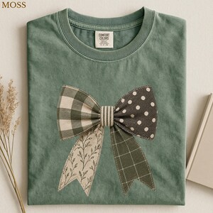 May include: A moss green crew neck t-shirt featuring a large central graphic of a bow. The bow is composed of four distinct fabric patterns: green and white gingham, white polka dots on dark green, a leafy vine print, and a green plaid pattern.