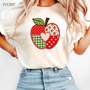 May include: An ivory t-shirt featuring a large graphic of a patchwork apple. The apple design is composed of four distinct sections with red polka dots, green and white gingham, red floral patterns, and red with white hearts, plus a central heart.