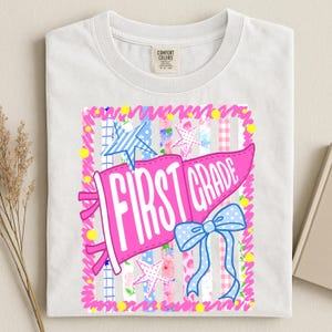 May include: A white t-shirt with a Comfort Colors label featuring a graphic of a pink pennant flag with the text FIRST GRADE in white. The design includes a blue polka dot bow, blue and white stars, and a background of vertical stripes.