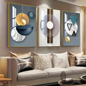 May include: Three framed abstract art pieces with gold frames. Each features geometric shapes in gold, blue, white, and gray. The artwork hangs above a light gray sofa with decorative pillows.
