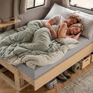 Puede incluir: Una pareja durmiendo en una autocaravana. Están acostados en una cama con un edredón verde claro y sábanas grises. La cama es de madera clara. Hay almohadas, una pequeña mesa de madera y una alfombra en la furgoneta.