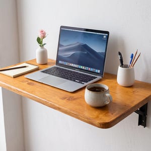 Puede incluir: Un escritorio flotante de madera montado en la pared con acabado natural, que incluye un portátil plateado, un pequeño jarrón blanco con una rosa rosa, un cuaderno de espiral con un lápiz, una taza de cerámica y un portalápices blanco.