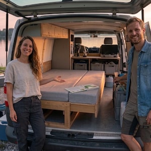 Op de afbeelding: Een camperinterieur met een bed, opbergruimte en een kleine keuken. Twee mensen staan voor de open achterkant van de bus, glimlachend. De scène speelt zich af in de buurt van een meer bij zonsondergang.