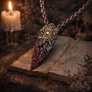 Puede incluir: Un collar con colgante de estilo gótico con una piedra preciosa roja y dorada. El colgante tiene un aspecto texturizado y dentado con una combinación de colores rojo y gris. El collar se muestra sobre un libro abierto, con una vela encendida en el fondo.