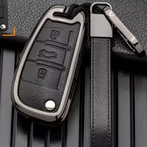 Può includere: Una custodia nera per chiave auto con cornice in argento metallizzato e cinturino in pelle nera abbinato. La custodia presenta tre pulsanti in rilievo con icone di blocco, sblocco e bagagliaio, con un piccolo rivetto color argento.