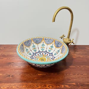 Könnte beinhalten: Ein rundes Keramik-Aufsatzwaschbecken mit handbemaltem, komplexem Blumen- und Geometriemuster in Blau-, Gelb-, Orange- und Türkistönen. Das Becken hat einen hellblauen Rand und einen hohen, geschwungenen Wasserhahn in antikem Messing-Finish.