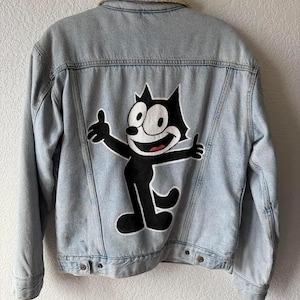 May include: Light blue denim jacket with a cream-colored sherpa collar. The back of the jacket features a large black and white graphic of Felix the Cat with his arms outstretched. The jacket is hanging on a metal hanger.