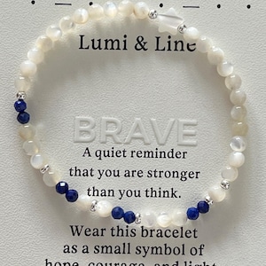 May include: A delicate bracelet featuring white beads and small blue beads, with silver accents. The text "Lumi & Line" and "BRAVE" are visible. The bracelet is a symbol of hope, courage, and light.