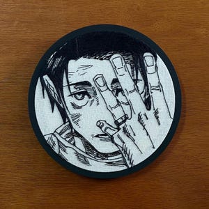 May include: Round embroidered patch featuring a black and white illustration of a person covering their face with their hand. The patch has a black border and is on a wooden surface. The illustration is detailed with visible facial features and a ring on one finger.
