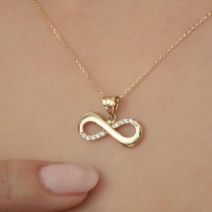Può includere: Una collana con ciondolo a infinito in oro composta da una catenina sottile e un simbolo dell'infinito in oro lucido decorato con piccole pietre chiare e scintillanti incastonate lungo un lato dell'anello, su un tessuto nero testurizzato.