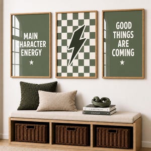 May include: Three framed wall art prints with light wood frames. The left print is olive green with the white text MAIN CHARACTER ENERGY and a star. The centre print features a green and white checkerboard pattern with a lightning bolt. The right print is olive green with the white text GOOD THINGS ARE COMING and a star.