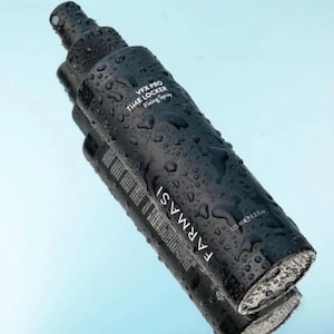 Pode incluir: Frasco preto de spray fixador Farmasi VFX Pro Time Locker, com gotas de água. O frasco contém 125 ml de produto. O frasco está sobre uma superfície refletora com fundo azul claro.