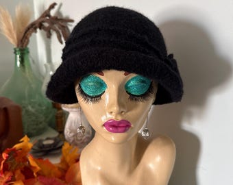 May include: A black cloche hat with a textured, fuzzy surface and a subtle ribbed design around the crown. The hat features a small decorative bow detail on the side and is displayed on a mannequin head with bright teal eyeshadow and pink lips.
