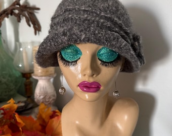May include: A dark grey, textured, knit cloche hat with a rounded crown and a slightly flared brim. The hat features a subtle, horizontal ribbed pattern around the crown and a decorative fabric detail on the side, providing a structured shape.