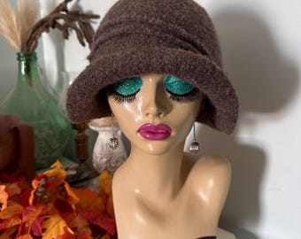May include: A brown felted cloche hat with a textured surface and a slightly downturned brim. The hat features a rounded crown with subtle horizontal pleating details around the base, providing a structured shape that sits atop a mannequin head.
