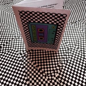 May include: A small white booklet with a black and white chequered border and a central illustration of a teal cassette tape labelled I Love the 80s Tarot. The interior pages show printed text including the words connection, endings, beginnings, and progress.