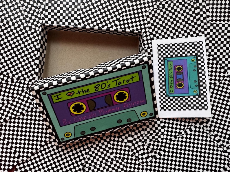 May include: A tarot card deck box and a small booklet featuring a black and white chequerboard pattern. The box lid displays a colourful illustration of a cassette tape with the text I Love the 80's Tarot by Christy Plummer Reynosa.