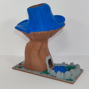 May include: A whimsical, fantasy-themed sculpture featuring a brown tree-like structure with a blue canopy. The base includes a small cave entrance and a miniature pond with blue water and green accents. The sculpture is set on a rectangular base.