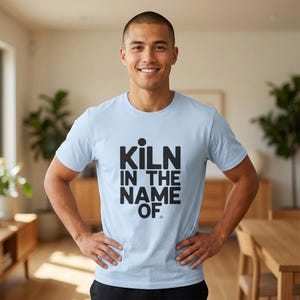 May include: A pale blue t-shirt featuring the words "KILN IN THE NAME OF" in black, bold font. The person wearing the shirt has their hands on their hips. The shirt is short-sleeved.