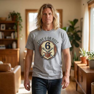 May include: A gray t-shirt with the text "GONE 6 OR BUST" encircling a shield-shaped graphic. The graphic features the number 6, crossed tools, and a brown and red color scheme. The shirt is worn by a person in blue jeans.