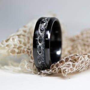 May include: A black band ring featuring a central inlay with a textured, dark grey, scale-like pattern. The ring has a polished finish on the edges and a smooth, rounded profile, showcasing a distinct contrast between the dark metal and the inlay.