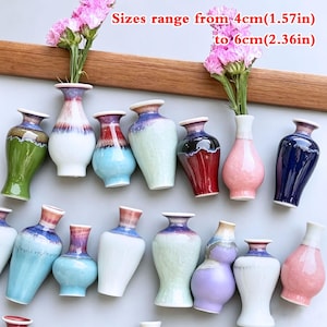 Puede incluir: Una colección de pequeños jarrones de cerámica en varios colores, incluyendo verde, azul, rojo, rosa y morado, con acabados esmaltados y cuellos estrechos. El texto indica tamaños de 4cm a 6cm. Algunos jarrones contienen flores rosas.